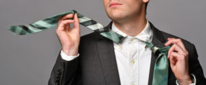 Ein junger Mann bindet sich schwungvoll seine Krawatte. Er trägt ein weißes Hemd, dunkles Sakko, die Krawatte ist schwarz, weiß und grün gemustert.
